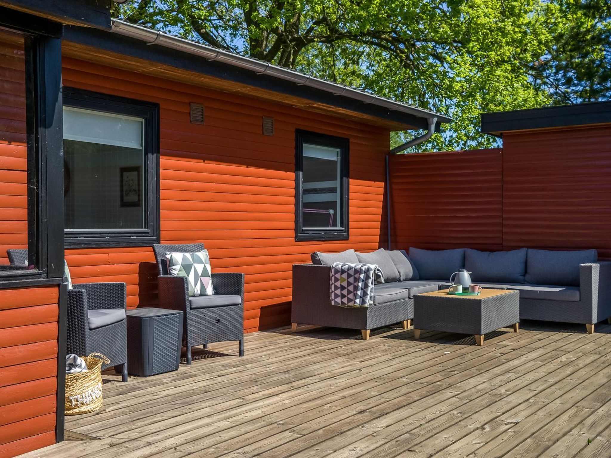 Une terrasse en bois avec des meubles d’extérieur, notamment des canapés et des fauteuils, et une fenêtre équipée de stores.