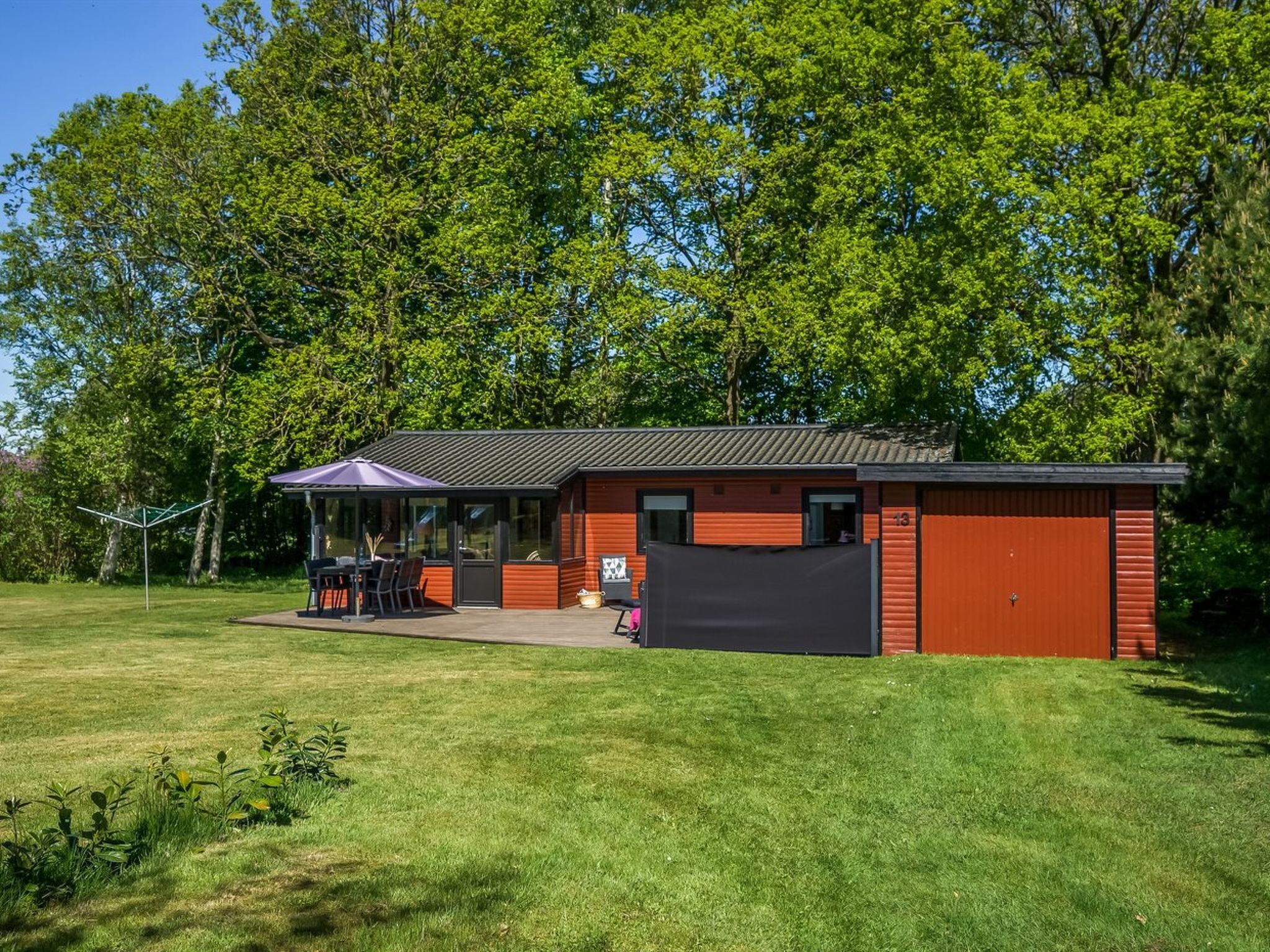Ein kleines, eingeschossiges Gebäude mit roter Fassade und einer schwarzen Garagentür. Es besitzt eine Terrassenfläche mit Gartenmöbeln und einen überdachten Sitzbereich.