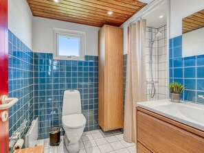 Une salle de bain avec des toilettes blanches, des carreaux bleus, un meuble en bois, un espace douche et une petite fenêtre.