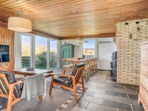 A cozy interior with a wooden ceiling, a dining area with chairs and a table, a kitchen with a brick wall, and large windows offering a view of the outdoors.