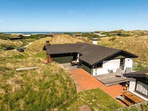Une maison moderne avec un toit en pente, une terrasse dotée de mobilier d’extérieur et une vue sur la plage.