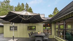Une terrasse extérieure avec une table et des chaises sous un grand parasol, entourée de murs et de fenêtres verdoyants.