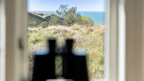Une paire de jumelles est posée sur un rebord de fenêtre, offrant une vue sur un paysage côtier avec de la végétation et un bâtiment.