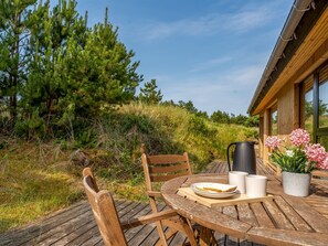Une terrasse extérieure en bois, avec une table dressée pour deux personnes, entourée de verdure et bordée d’un bâtiment doté de grandes fenêtres.