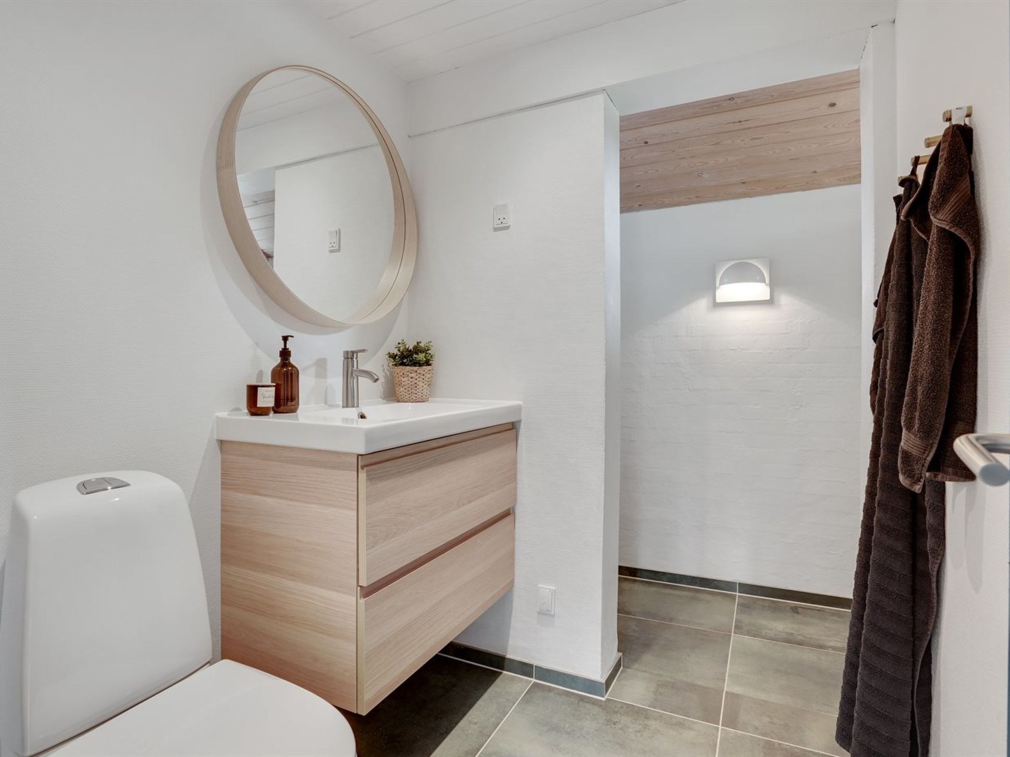 Un baño moderno con un lavamanos blanco, tocador de madera, un espejo redondo y un perchero para toallas.