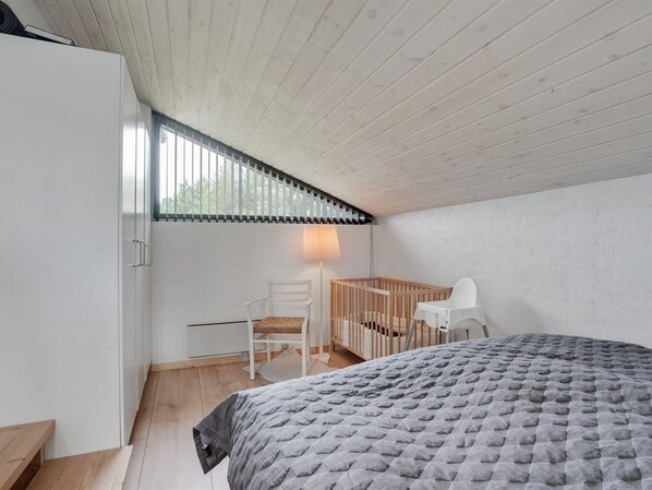 A bedroom with a wooden ceiling, a bed with a grey quilt, a crib, a white chair, and a window with blinds. - "Kadife" - 750m from the sea by Interhome (Saltum)