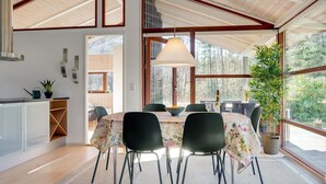 Un coin repas avec une table ronde, des chaises noires et une nappe à motifs floraux. Une grande fenêtre offre une vue sur les arbres, et un luminaire suspendu éclaire l’espace.