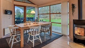 A cozy cabin interior with a wooden dining table, chairs, a large window, and a wood-burning stove.