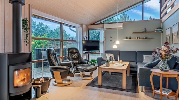 A modern living room with a wood-burning stove, leather chairs, a sofa, and large windows offering a view of the outdoors.