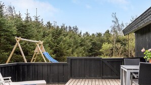 Een houten terras met een schommel en een glijbaan, omringd door een houten hek en weelderige bomen.
