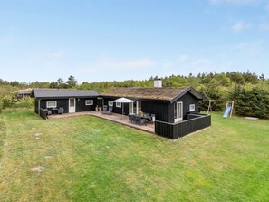 Eine schwarze Holzhütte mit einer Terrasse und einer Rutsche, umgeben von einem grünen Rasen und Bäumen.
