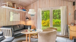 A modern living room with a black leather sofa, a wooden coffee table, and a beige armchair. There is a bookshelf with books and a ship model. A large window with curtains offers a view of greenery outside.