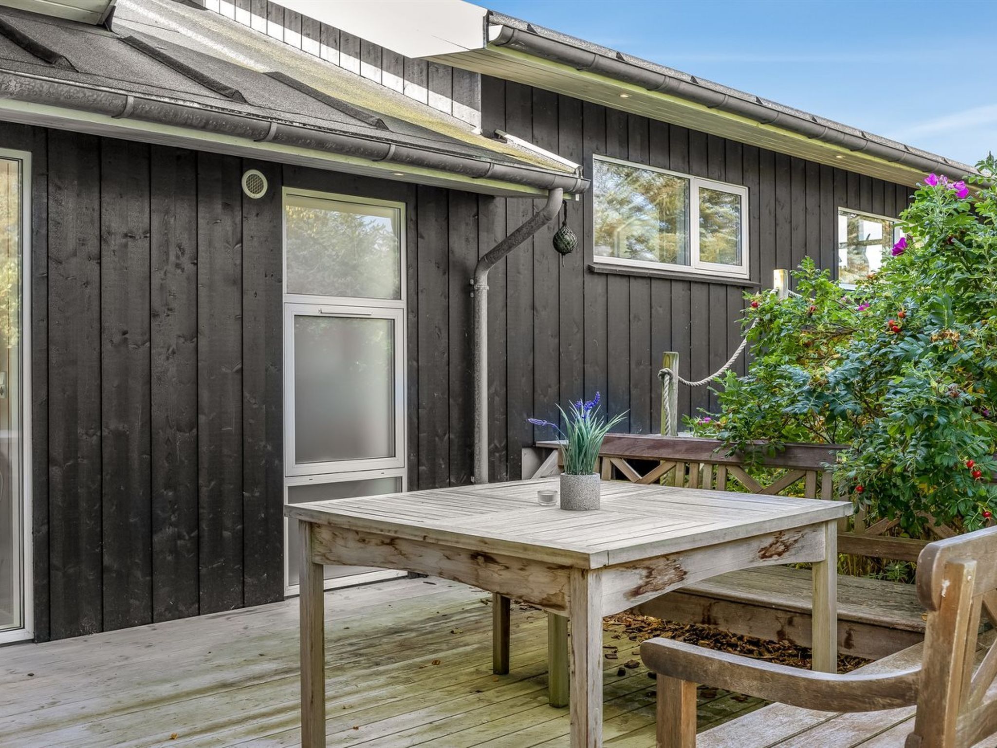 Een houten terras met een tafel, stoelen, een plant in pot en een tuin met bloemen en een boom.
