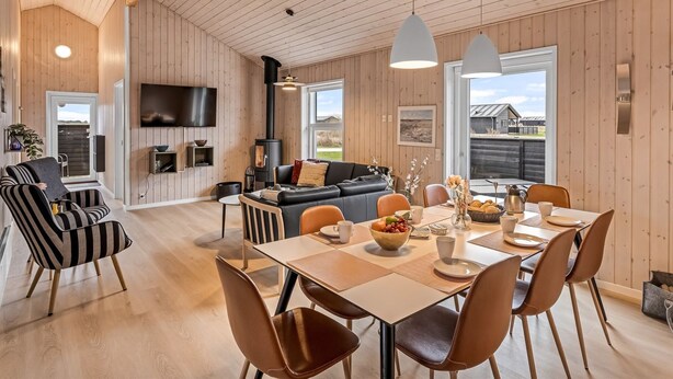 A spacious living area with a wooden ceiling, a black sofa, a dining table with chairs, and a television mounted on the wall.