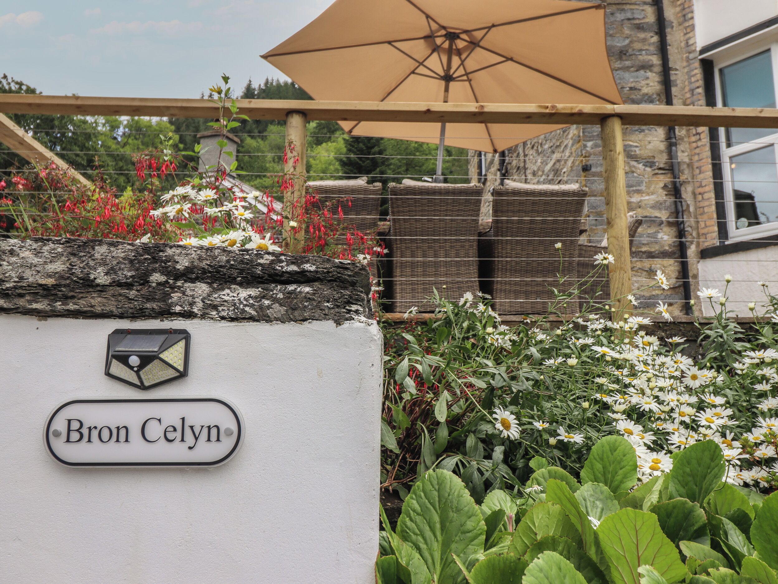 A sign with the name 'Bron Celyn' on a white wall, a wooden pergola with a beige umbrella, and a garden with various plants and flowers.