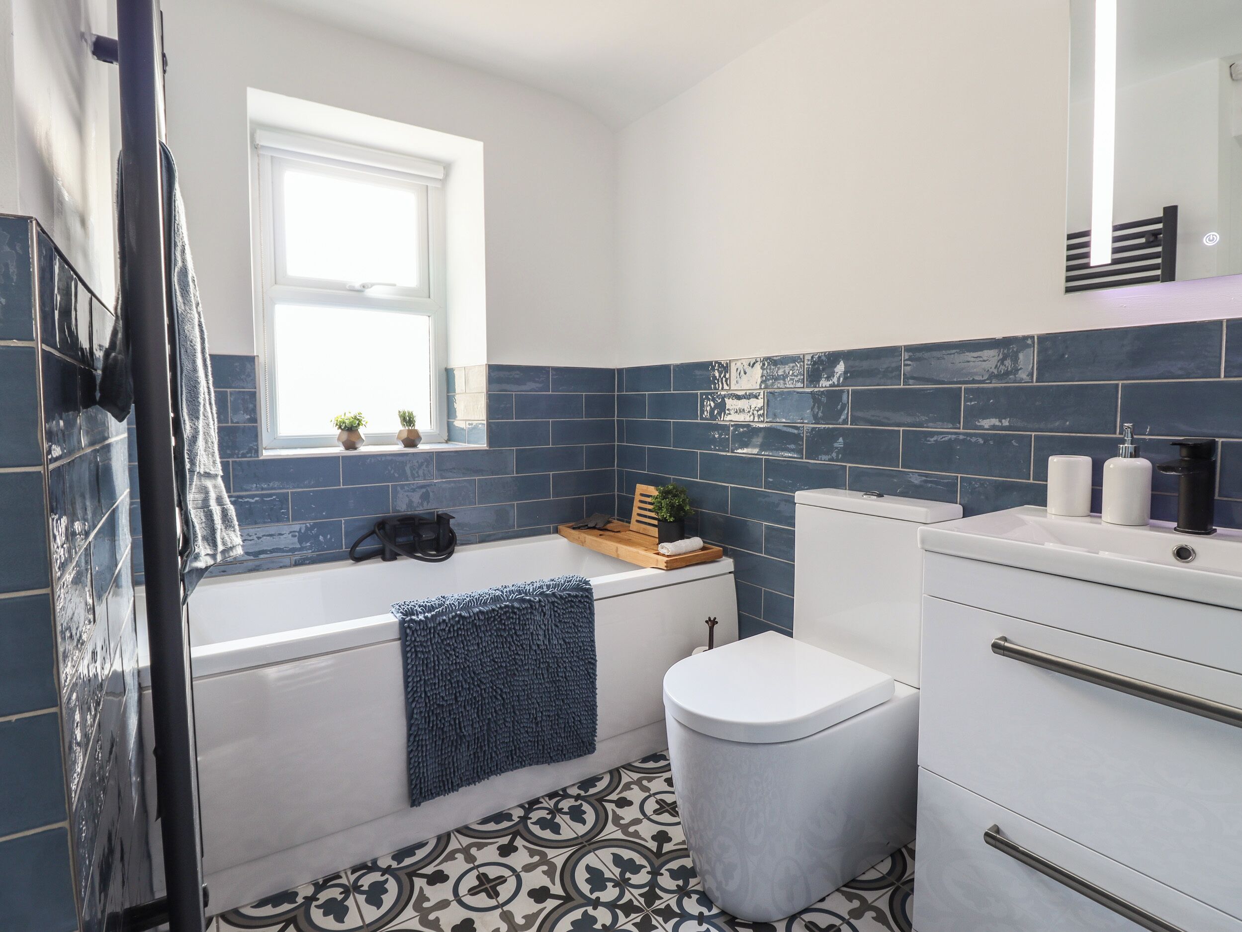 Un baño moderno con bañera, inodoro y lavamanos. Hay un ventanal sobre la bañera y un espejo sobre el lavamanos.