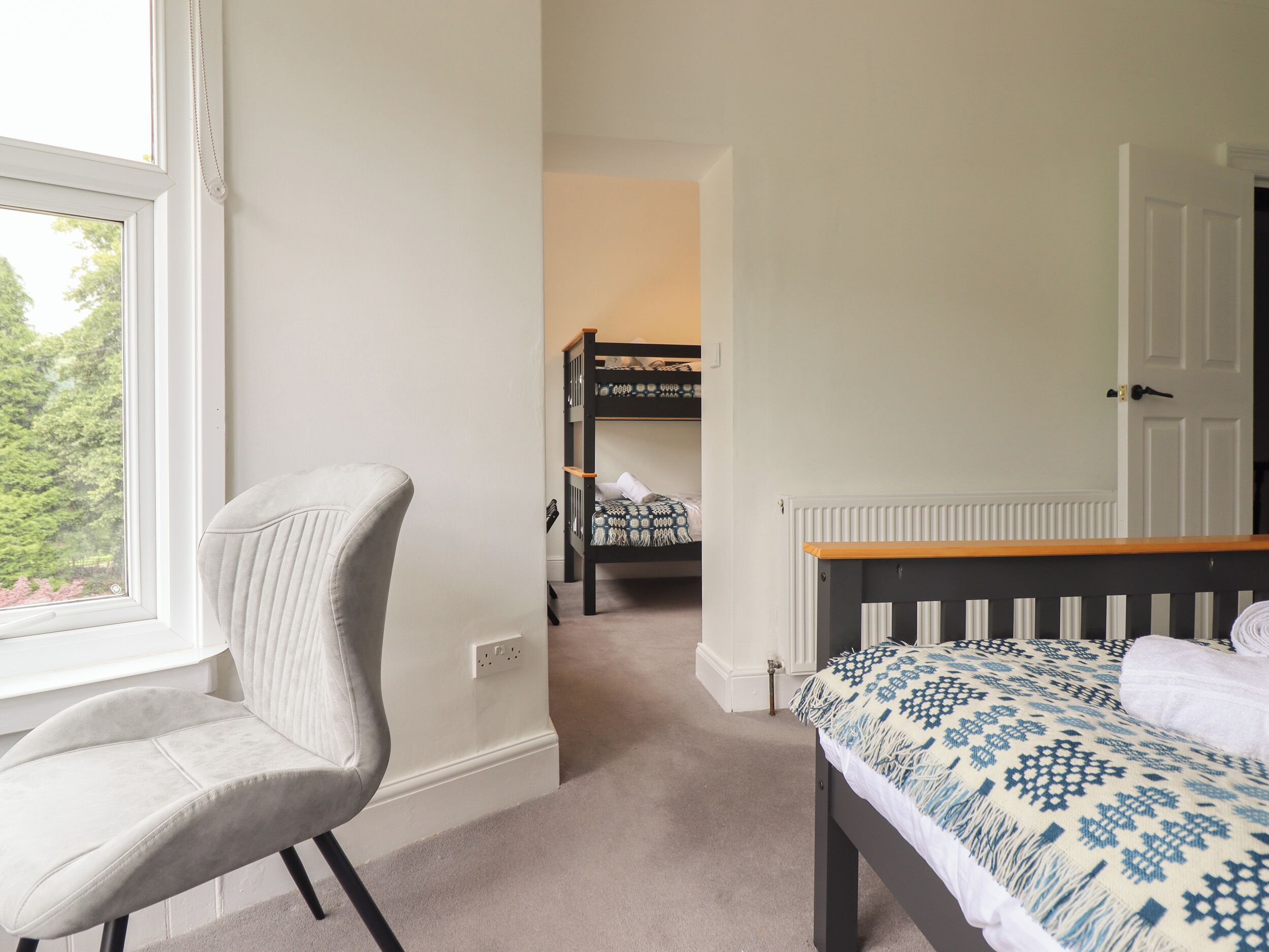 A bedroom with a single bed, a chair, a bunk bed in the background, and a window with a view of trees.