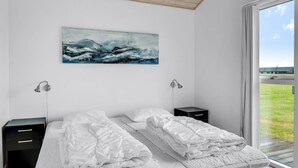Un dormitorio moderno con dos camas, un cuadro grande de un paisaje marino y vista a un área verde a través de un amplio ventanal.