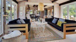 A spacious living area with a large sofa, a coffee table, and a dining table. There are multiple chairs and a hanging light fixture.