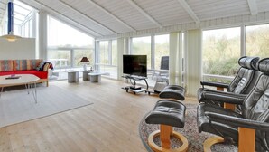 Ein geräumiges Wohnzimmer mit hoher Decke, großen Fenstern, einem Sofa, einem Couchtisch, einem Fernseher und Ledersesseln.