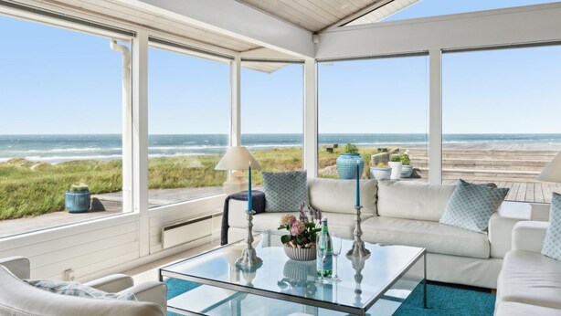 Una sala de estar con ventanales, una mesa de centro de vidrio, sofás blancos y vista a la playa.