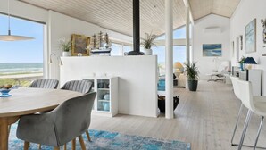 Amplia sala de estar con techo alto, un ventanal con vista al océano, chimenea central y una cocina moderna.