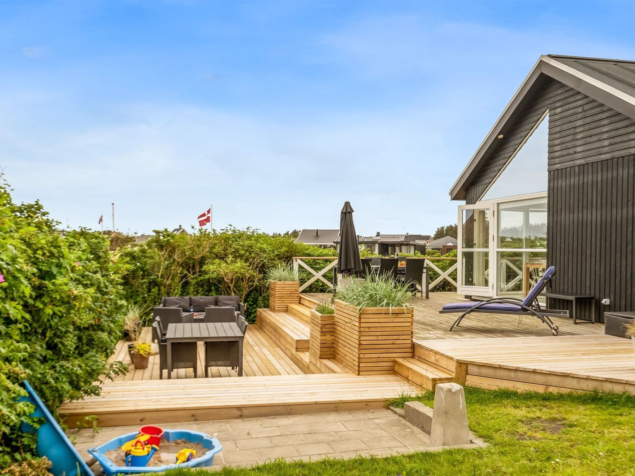 Een modern terras buiten met een houten tafel en stoelen, een klein speelgedeelte met een glijbaan en zandbak, en een ruime kamer met ramen.