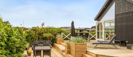 Een modern terras buiten met een houten tafel en stoelen, een klein speelgedeelte met een glijbaan en zandbak, en een ruime kamer met ramen.