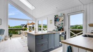 Une cuisine moderne dotée d’un grand îlot central, de murs en bois peints en blanc et de grandes fenêtres offrant une vue sur l’extérieur.