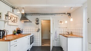 Una cocina moderna con armarios blancos, encimera de madera, estufa y fregadero. Hay ollas y sartenes colgando, y una cafetera sobre la encimera.