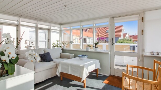 Una habitación luminosa e soleada con un sofá blanco, una mesita con mantel blanco y una silla de madera.