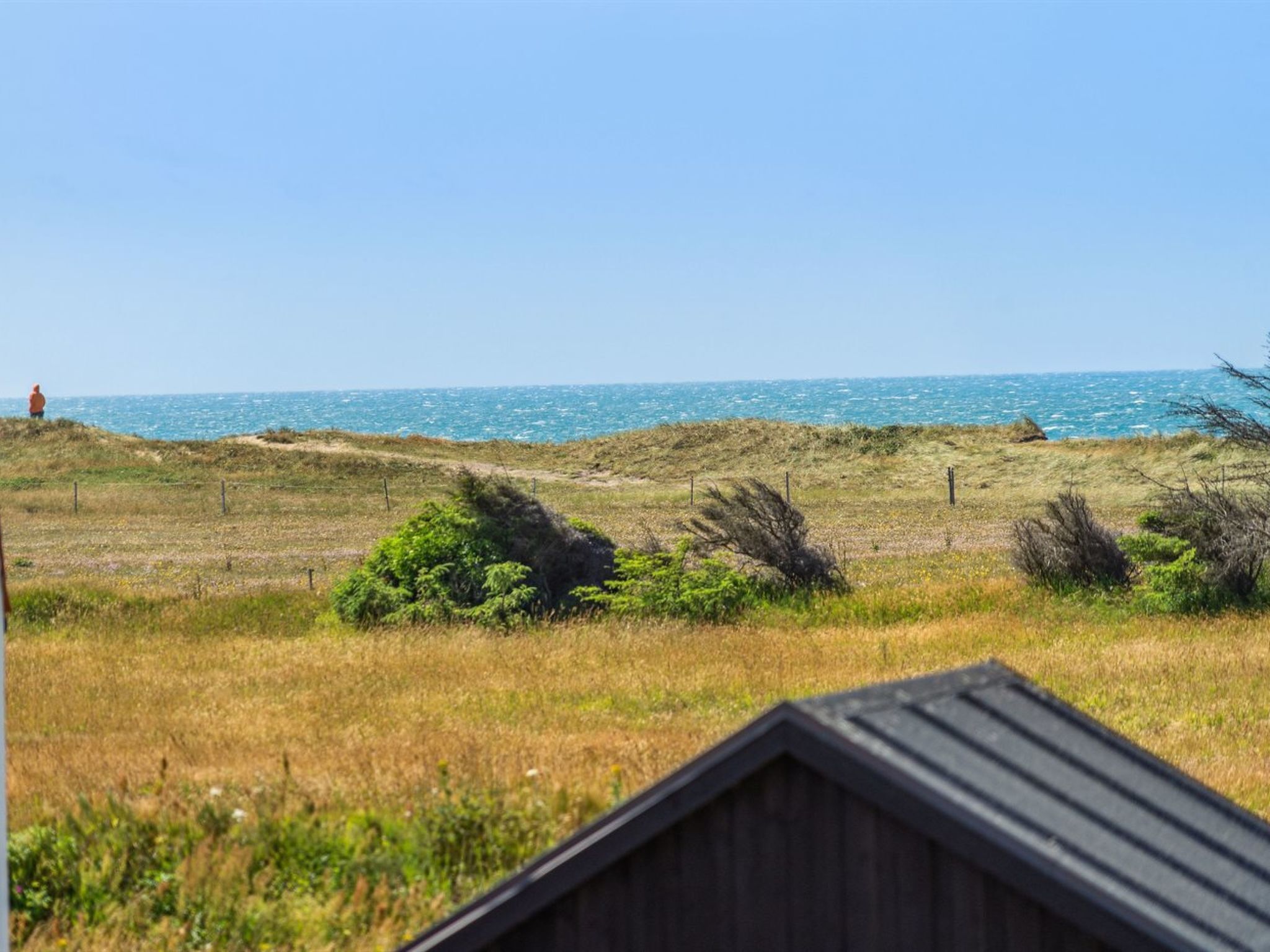 A coastal landscape with a clear view of the sea, a grassy area with sparse vegetation, and a wooden structure in the foreground.