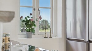 A kitchen with a window, a potted plant, a candle, a mug, a bowl of fruit, and a refrigerator.