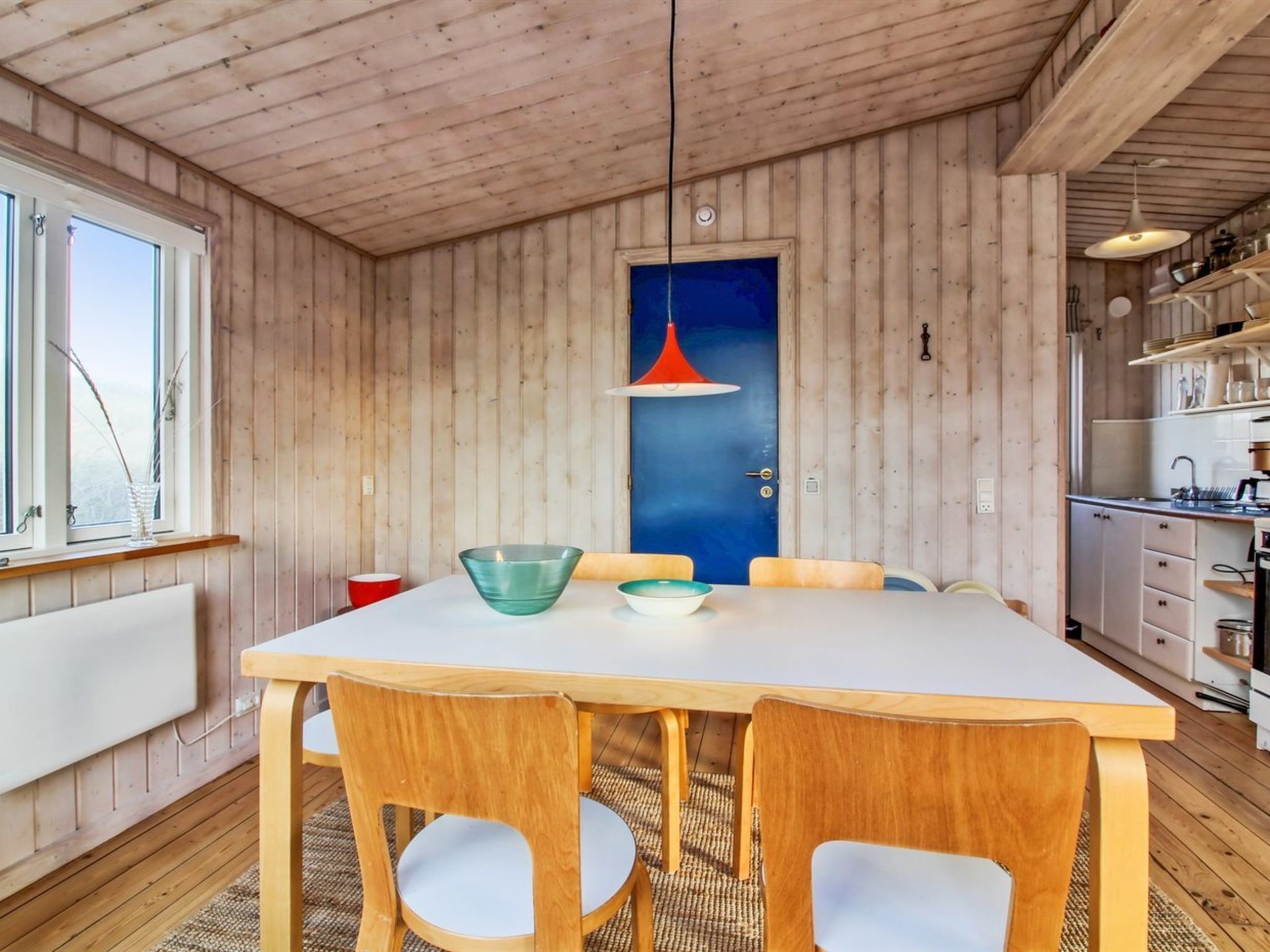 Une pièce aux murs lambrissés, avec une grande table, des chaises, une porte bleue et un coin cuisine comprenant des placards et un évier.