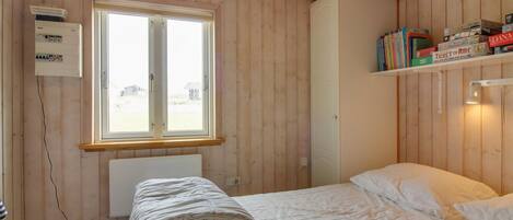Una habitación con paredes de madera, una cama con ropa de cama blanca, una ventana con vistas, y una estantería con libros y un juego de 'Ticket to Ride'.