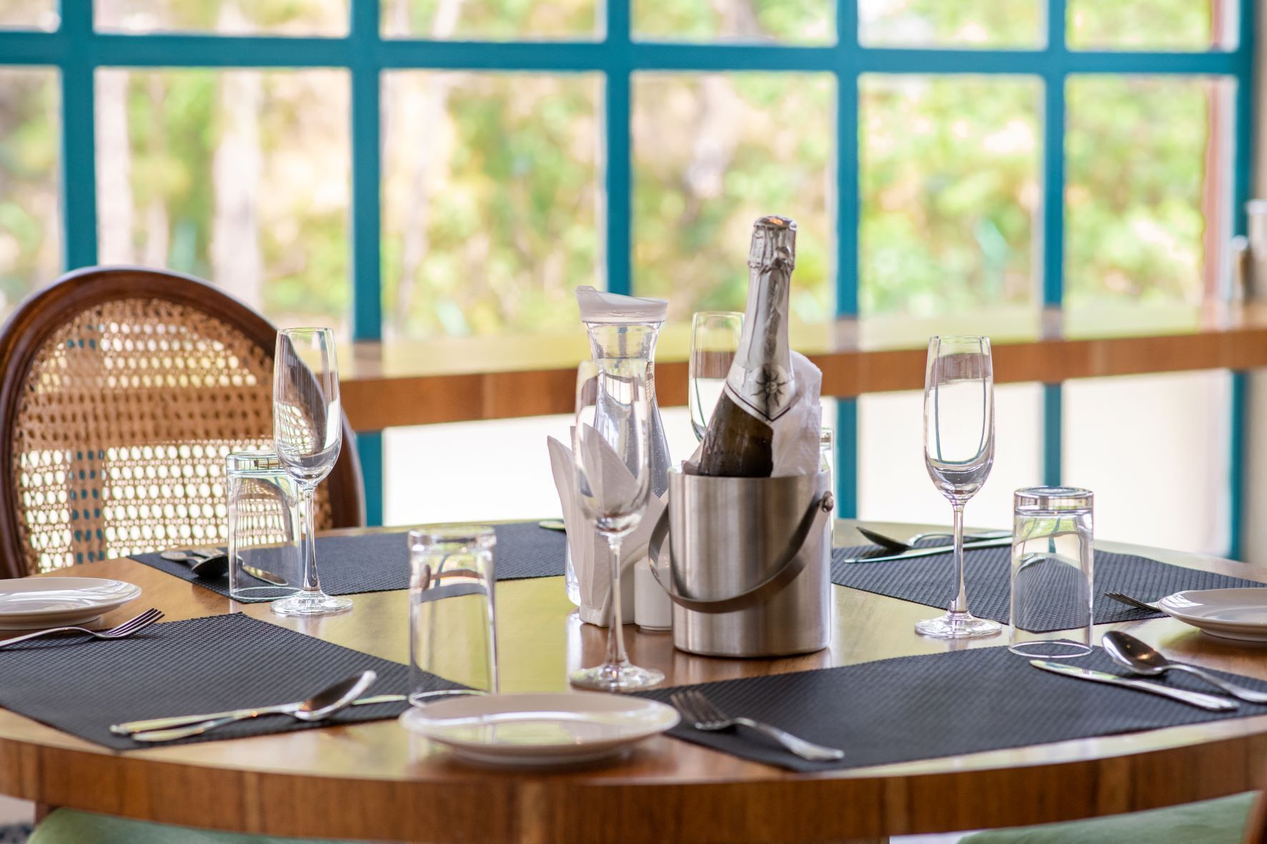 Una mesa de comedor dispuesta con copas de vino, una botella de champán en un cubo plateado y una silla de mimbre.