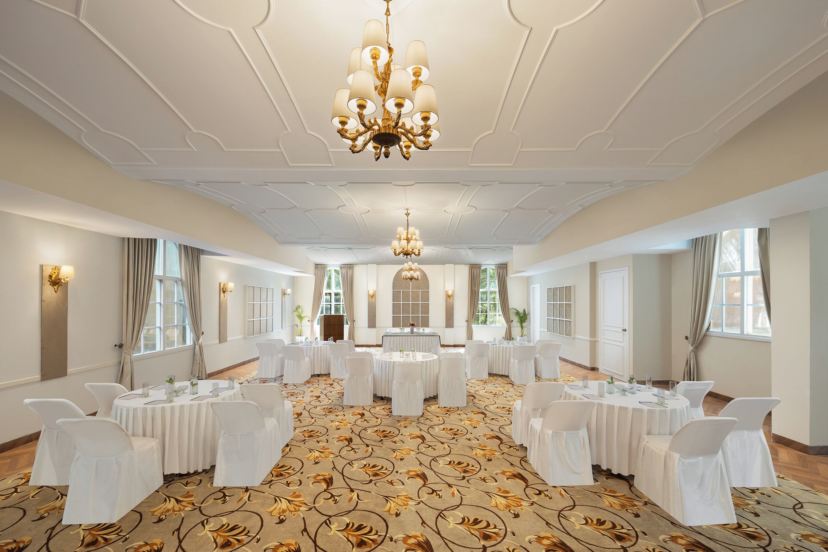 Une salle de banquet spacieuse, avec des tables rondes, des chaises blanches et un lustre central.