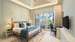 Habitación de hotel con una cama grande, un escritorio con silla, televisor y balcón con vistas a árboles.