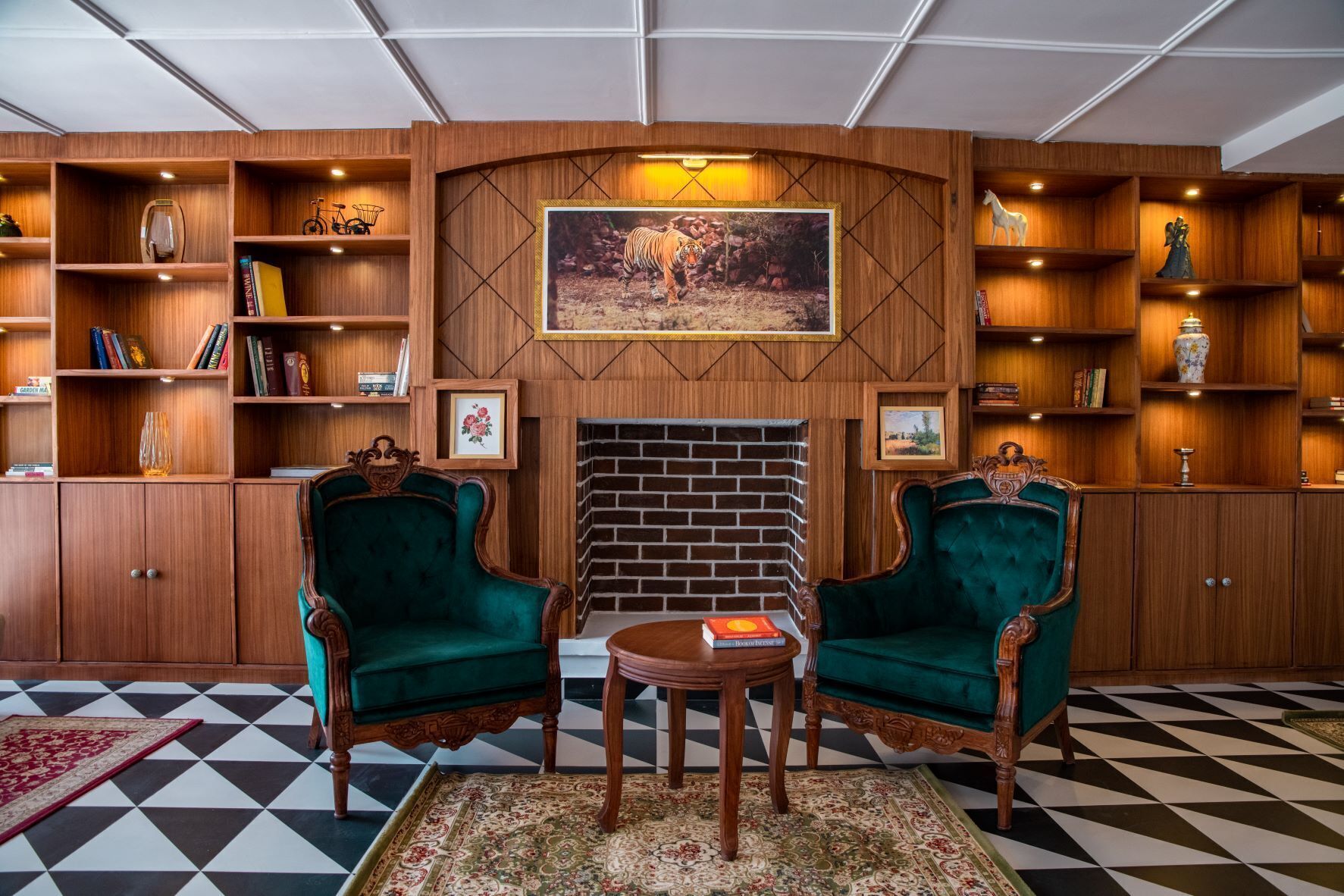 Um quarto com duas poltronas estofadas em verde, uma mesinha redonda, lareira e estantes de madeira com livros e objetos decorativos.