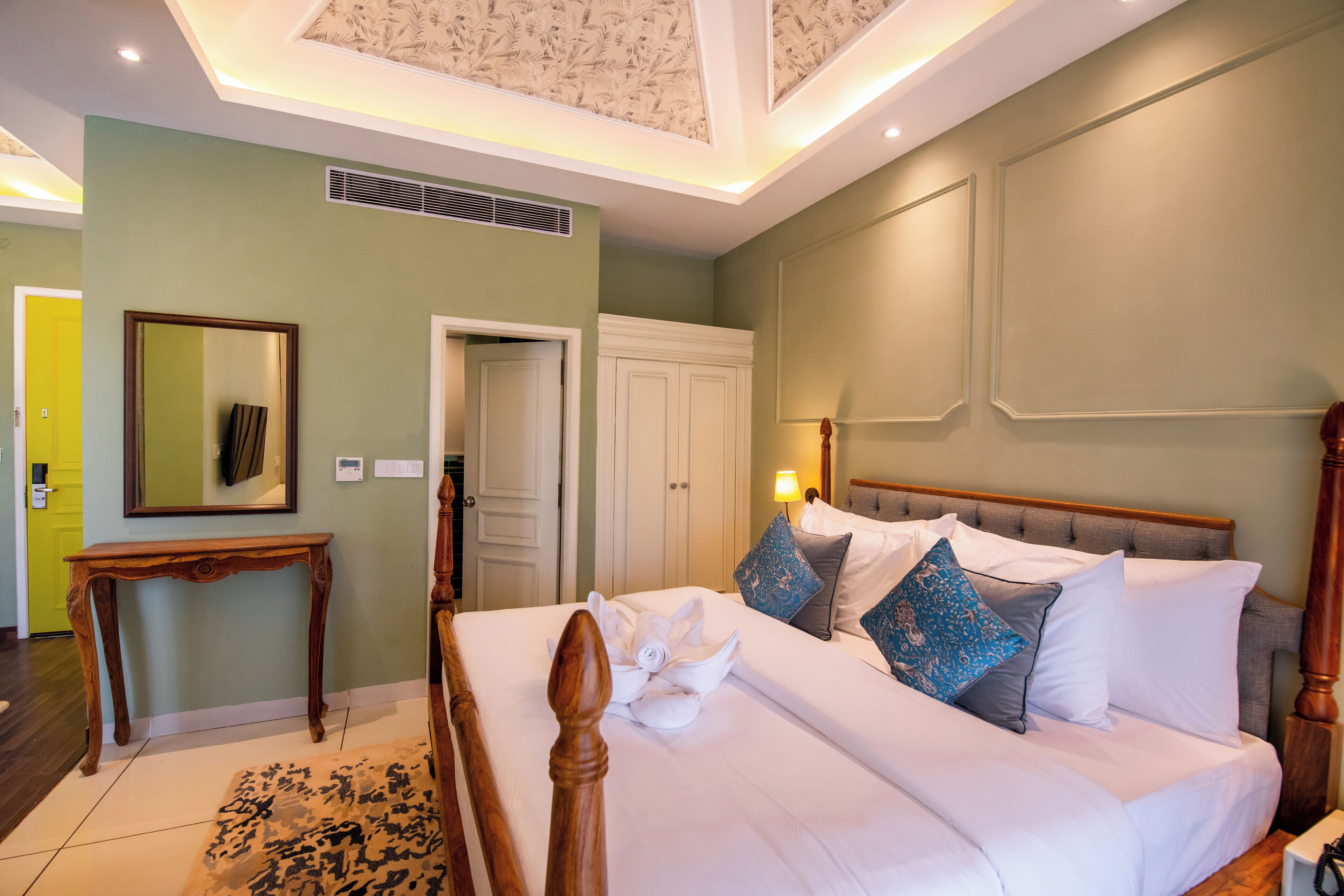 Una cama bien hecha con sábanas blancas y almohadones decorativos, una mesita de noche de madera y un espejo en la pared.