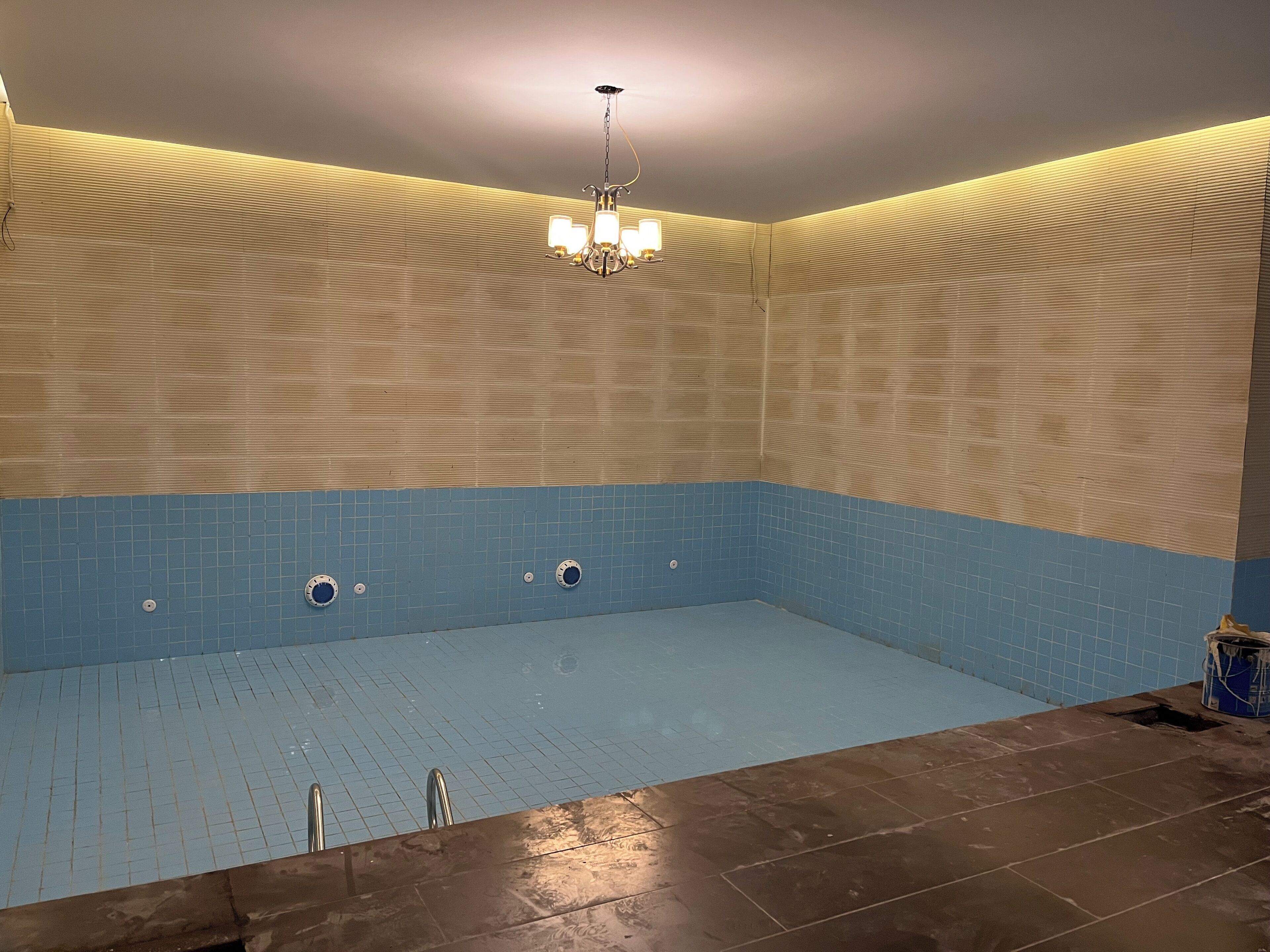 Une piscine dont l’intérieur est revêtu de carreaux bleus, ornée d’un lustre et dont un des murs est également revêtu de carreaux présentant une texture à motifs en relief.