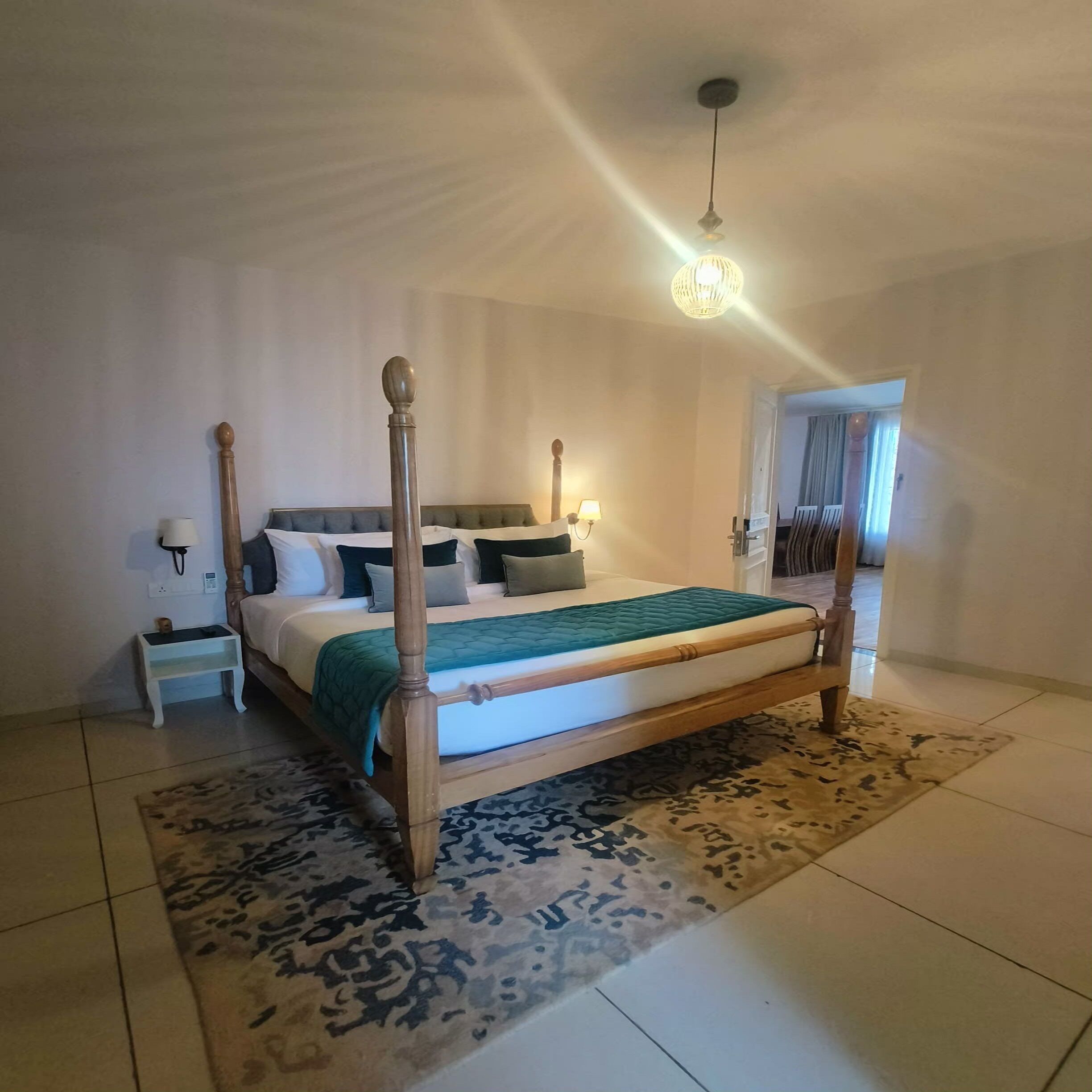 Uma cama de dossel com estrutura de madeira, colcha verde e tapete estampado. Há uma mesa de cabeceira com abajur e telefone. O quarto tem paredes brancas e uma luminária no teto.