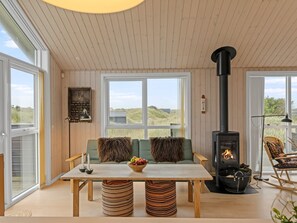 Un intérieur chaleureux avec un canapé, une table sur laquelle est posé un panier de fruits, une cheminée et de grandes fenêtres donnant sur l’extérieur.
