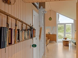Une pièce avec une bibliothèque remplie de livres, une structure en poutres apparentes, une porte blanche et une vue sur un espace extérieur aménagé avec une table et des chaises.
