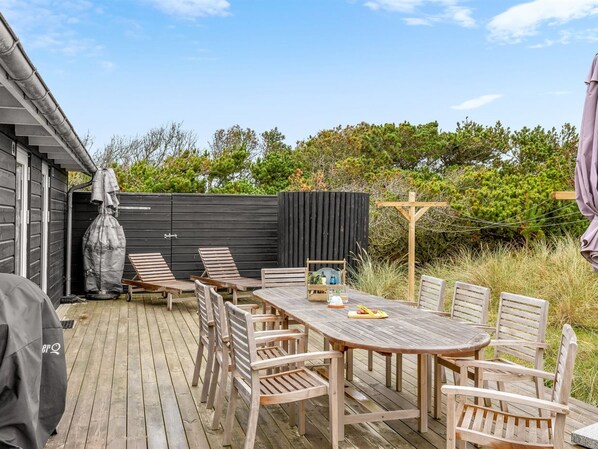 Una terraza exterior de madera con una mesa, sillas, una parrilla y un área cubierta con un banco.