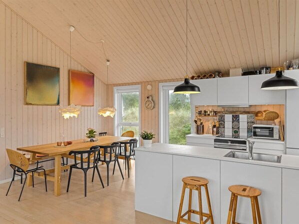 Una cocina moderna con armarios blancos, una isla central con taburetes, iluminación colgante y un comedor con mesa y sillas de madera.