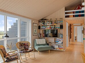 Una habitación acogedora con un ventanal grande, una estantería llena de juegos de mesa, un televisor y dos mecedoras.