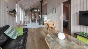 Ein gemütliches Cabin-Interieur mit Holzbalkendecke, einer schwarzen Ledersofa, einer Essecke mit Korbstühlen, einem Kamin und einem Fernseher.