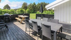 Eine Terrasse im Freien mit Holzdeck, Tisch und Stühlen, Grill und einem überdachten Bereich mit weißer Wand.