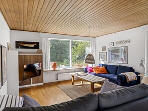 A modern living room with a wooden ceiling, a large window, a blue sofa, a wooden coffee table, and a television.